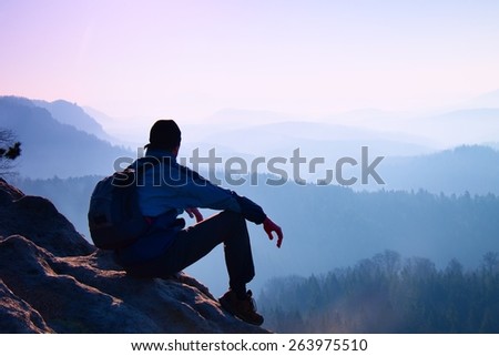 Tired hiker with sporty backpack sit on rocky peak  and watching into deep misty valley bellow. Sunny spring daybreak in rocky mountains.
