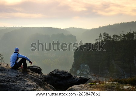 Moment of loneliness. Man sit on the peak of rock and watching into colorful mist and fog in forest valley.