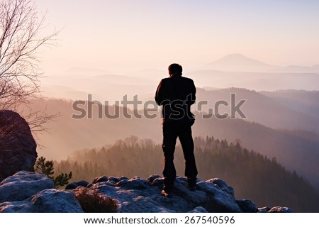 Professional on location.  Nature photographer  takes photos with mirror camera on peak of rock. Dreamy fogy landscape, spring orange pink misty sunrise in a beautiful valley below.
