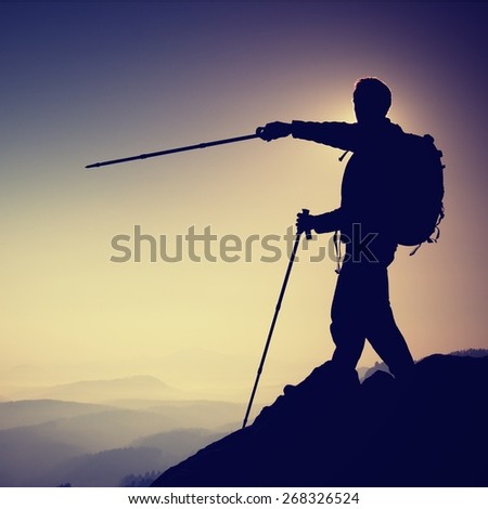 Tourist guide show the right way with pole in hand. Hiker with sporty backpack stand on rocky view point above misty valley. Sunny spring daybreak in rocky mountains.