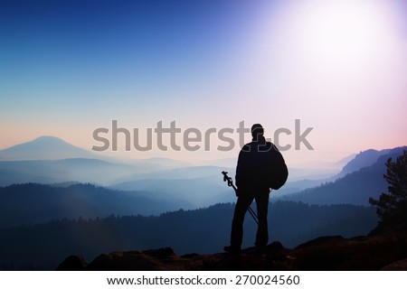 Silhouette of tourist  with poles in hand. Hiker with sporty backpack stand on rocky view point above misty valley. Sunny spring daybreak in rocky mountains.