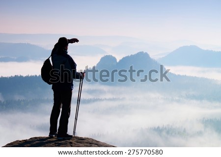 Tourist with backpack and poles in hand. Sunny spring daybreak in rocky mountains. Hiker with sporty backpack stand on rocky view point above misty valley.