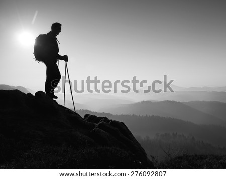 Tourist with sporty backpack and poles in hands on rock above valley. Sunny spring daybreak in rocky mountains. Black and white photo
