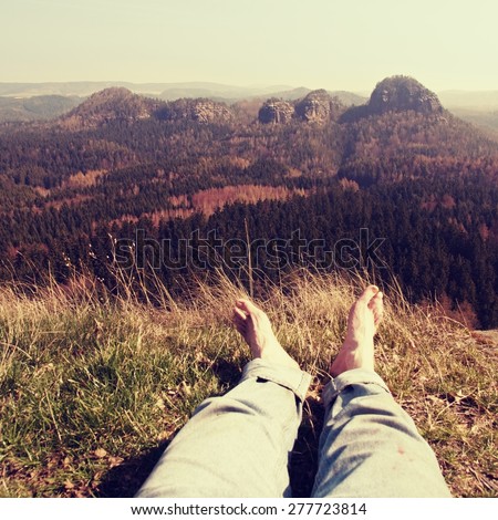 Naked male hairy legs in blue jeans take a rest on peak of rock above valley.