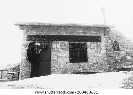 Tired tourist with backpack is knocking on locked door of stony mountain cottage. Winter in mountains. Black and white photo.