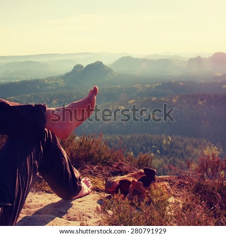 Naked male hairy legs in dark hiking trousers take a rest on peak of rock above spring valley.