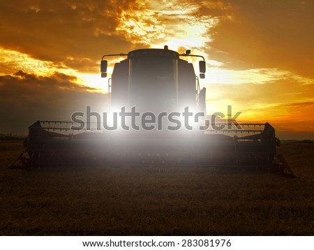 Abandoned combine harvest wheat with lighted main lights in the middle of a farm field. Morning yellow wheat field on the sunset cloudy orange sky background.