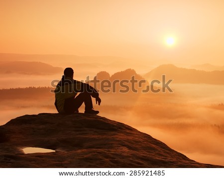 Hiker in black on the rocky peak. Wonderful daybreak in mountains, heavy orange mist in deep valley. Man sit on the rock and watch over the fog .