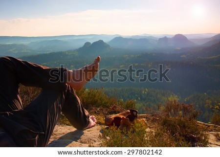 Naked male hairy legs in dark hiking trousers take a rest on peak of rock above spring valley.