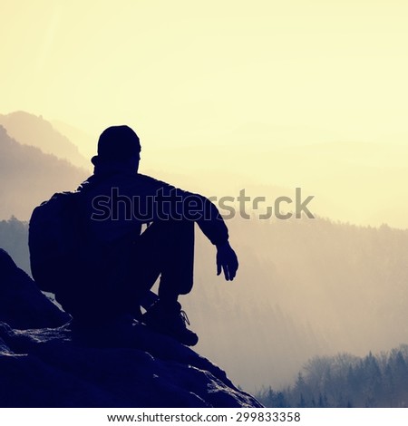 Tired hiker with sporty backpack sit on rocky peak and watching into deep misty valley bellow. Sunny spring daybreak