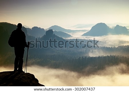 Silhouette of tourist with poles in hand. Sunny spring daybreak in rocky mountains. Hiker with sporty backpack stand on rocky view point above misty valley.