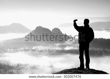 Tall tourist is taking selfie on peak above valley. Black and white photo