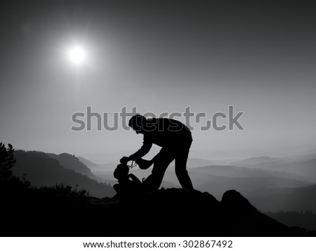 Professional photographer is packing camera into backpack on peak of rock. Dreamy fogy landscape, spring orange pink misty sunrise in a beautiful valley below. Black and white photo
