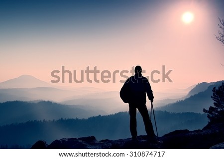 Silhouette of tourist with poles in hand. Hiker with sporty backpack stand on rocky view point above misty valley. Sunny spring daybreak in rocky mountains.