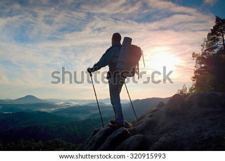 Tourist guide on the way with pole in hand. Hiker with sporty backpack stand on rocky view point above misty valley. Sunny spring daybreak in rocky mountains.