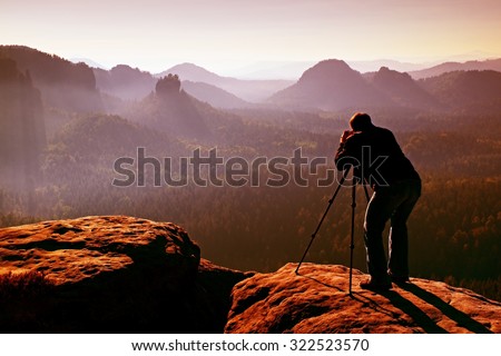Professional on cliff. Nature photographer takes photos with mirror camera on peak of rock. Dreamy fogy landscape, spring orange pink misty sunrise in a beautiful valley below.