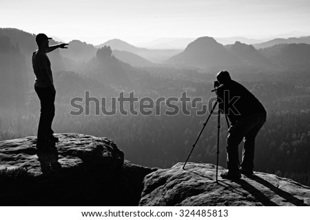 Hiker and photo enthusiast stay with tripod on cliff and thinking. Dreamy fogy landscape, blue misty sunrise in a beautiful valley below. Black and white photo