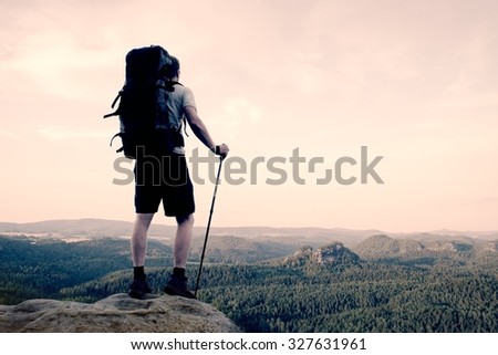 Tall tourist with poles in hand. Sunny evening in rocky mountains. Hiker with big backpack stand on rocky view point above misty valley.