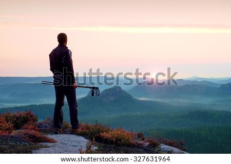 Silhouette of tourist guide with poles in hand. Hiker with sportswear stand on view point above misty valley. Sunny spring daybreak in rocky mountains.