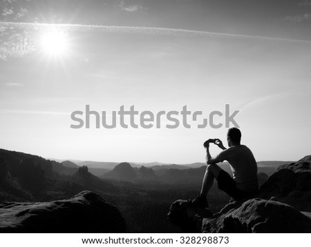 Tourist in grey t-shirt takes photos with smart phone on peak of rock. Dreamy hilly landscape below, spring misty sunrise . Black and white photo