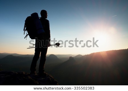 Tall backpacker with poles in hand. Sunny spring daybreak in rocky mountains. Hiker with big backpack stand on rocky view point above misty valley.