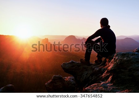 Adult tourist in black trousers, jacket and dark cap sit on cliff\'s edge and looking to misty hilly valley bellow