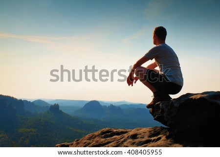 Young hiker in black pants and shirt  is sitting on cliff\'s edge and looking to misty hilly valley bellow