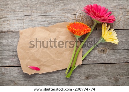 Three colorful gerbera flowers with paper for copy space on wooden table