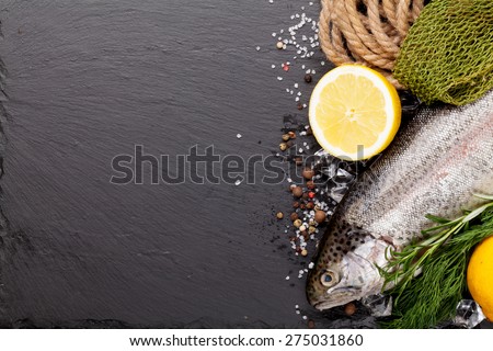 Fresh raw rainbow trout fish with spices and fishing equipment on black stone background. Top view with copy space