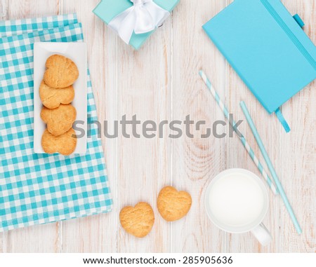 Cup of milk, heart shaped cookies, gift box and notepad on white wooden table with copy space