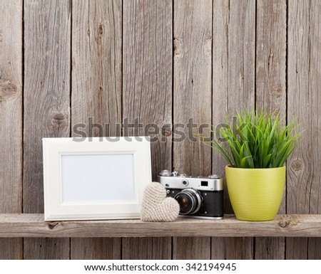 Blank photo frame, heart gift, camera and plant on shelf in front of wooden wall