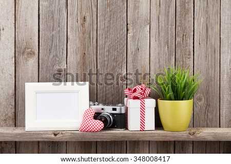 Blank photo frame, heart gift, camera and plant on shelf in front of wooden wall