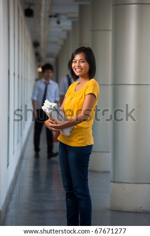 Three quarter length portrait of a cute laughing college student cradling books on a modern university campus.  Young female Asian Thai model late teens, early 20s of Chinese descent.