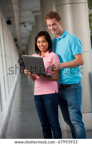 A pair of happy college students look up from sharing a laptop on a university campus.   20s female Asian Thai model of Chinese descent.  20s tall male caucasian model British nationality.