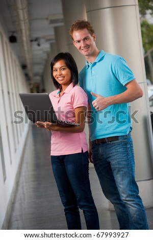 Two happy, smiling college students using a laptop at a gorgeous university campus hallway.  20s female Asian Thai model of Chinese descent.  Tall male caucasian model British nationality thumbs up.