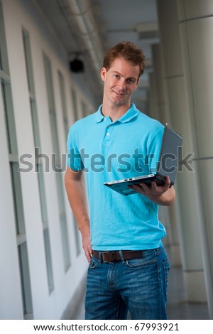 A confident college guy holds a laptop in a beautiful campus hallway.  20s tall handsome male caucasian British model.