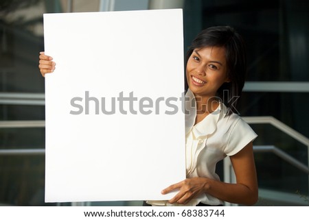 A friendly and pretty business professional holds a blank poster in front of an modern office complex.  Custom text insert.   20s female Asian Thai model of Chinese descent.