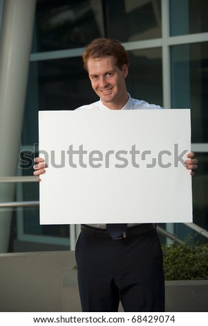 A happy and smiling office worker holding a blank sign outside of a glass office building.  20s handsome caucasian male British model.