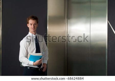 A happy and smiling office worker waiting at the elevator in a typical office building hallway.  20s handsome caucasian male British model.