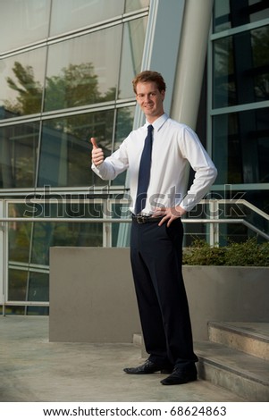 A happy corporate employee smiling and giving the thumbs up outside of a modern office park.  20s handsome caucasian male British model.
