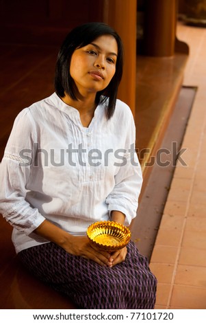 A pretty Thai woman in traditional clothes relaxes on the porch of a Thai style teak wood house.  20s female Asian Thai model of Chinese descent.