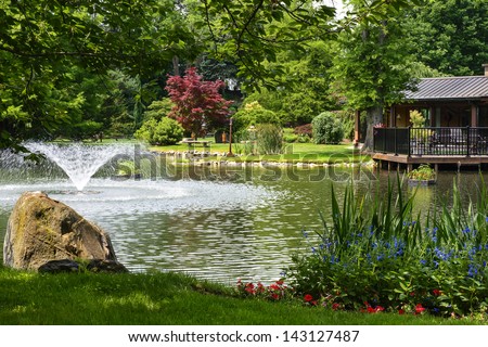 Beautiful Manicured Garden With A Deck Overlooking A Pond With A Fountain That'S Surrounded With Potted Plants And Flower Beds.