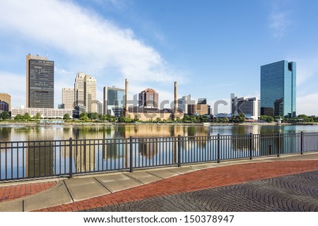 A panoramic view of downtown Toledo Ohio\'s skyline from across the Maumee river at a popular restaurant area with a paver brick boardwalk and a decorative iron railing..