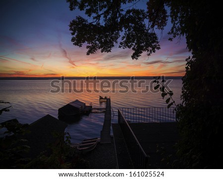 A beautiful sunrise on Lake Cayuga in the Finger lakes region of New York state. A row boat is docked on the side of a pier that leads out to a deck with chairs for watching the sunrise.
