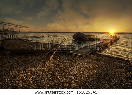 A beautiful sunrise on Lake Cayuga in the Finger lakes of New York state. A row boat is docked on the side of a pier that leads out to a deck with chairs for watching the sunrise.