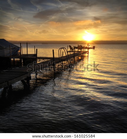 A beautiful sunrise on Lake Cayuga in the Finger lakes region of New York state. A  pier that leads out to a deck with chairs for watching the sunrise. Two fisherman are silhouetted in the sun.