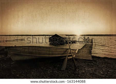 Photo of a lake shore dock with a row boat and boat shelter at sunrise. Photo has been enhanced with a sepia tone an textured for a grungy, vintage old photo  look.