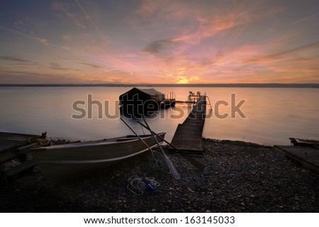 A beautiful autumn sunrise on Lake Cayuga in the Finger lakes region of New York state. A row boat sits on the side of a pier that leads out to a boat shelter and a deck for watching the sunrise.