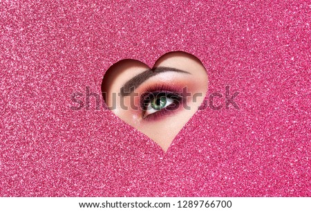 Conceptual photo of Valentine\'s day. Eye of Girl with Festive Pink Makeup. Paper heart on a pink background. Love symbols Valentines day