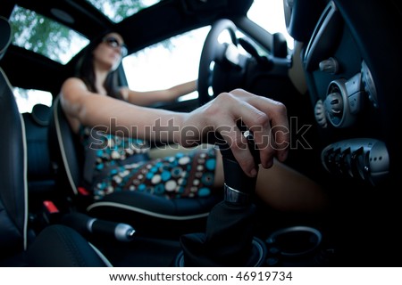 Woman driving, using a manual transmission stick shift. Photographed with  a fisheye lens for exaggerated perspective. Shallow depth of field with selective focus on hand.